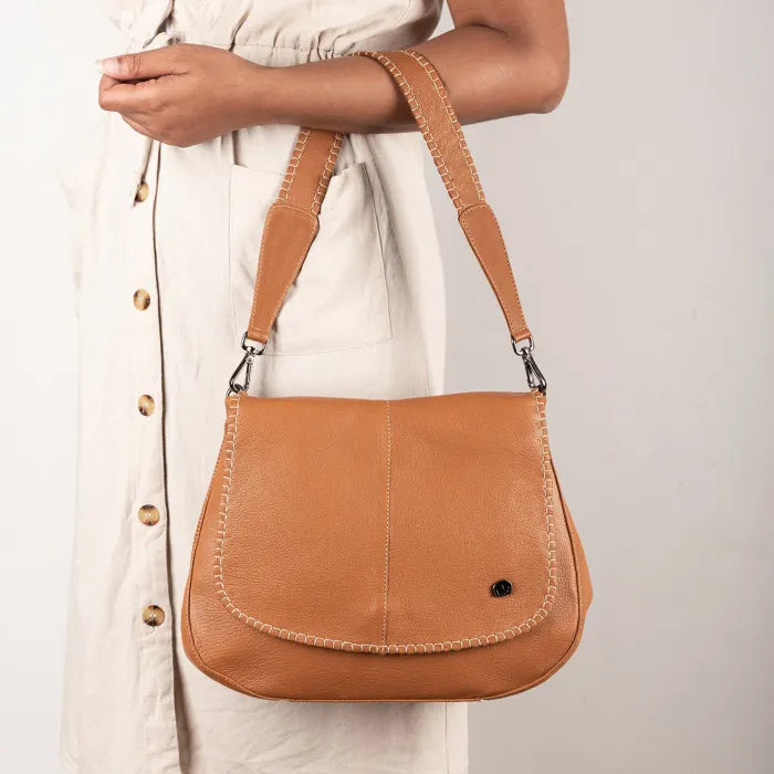Tan leather shoulder bag held by a person wearing a white dress.