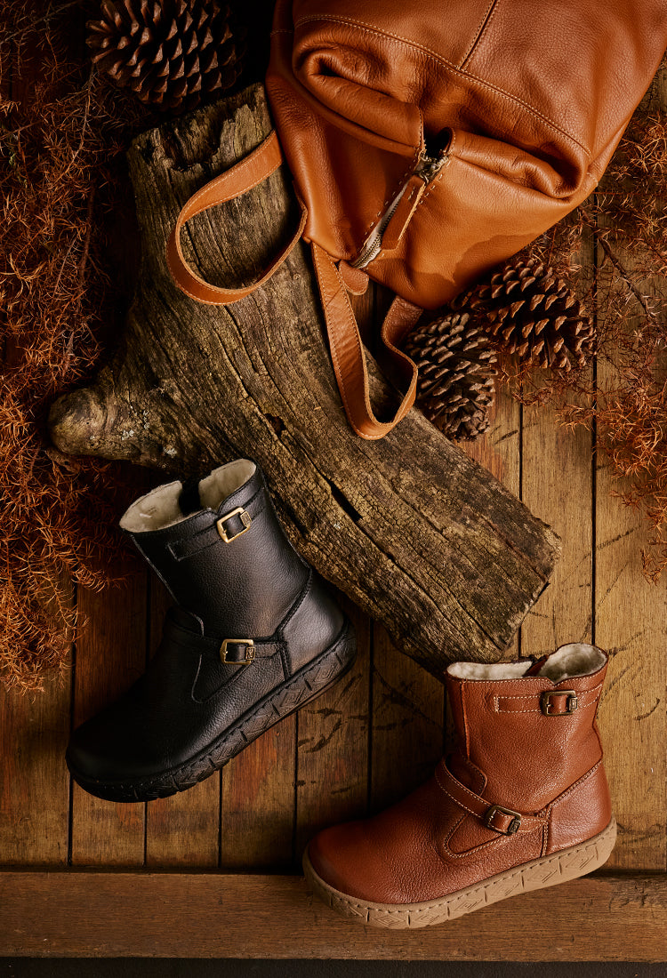 Pair of black and brown boots on a wooden surface with a leather bag and pinecones.