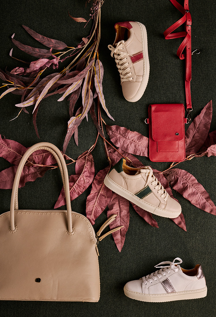 Beige handbag, white sneakers, and a red wallet on dark surface with pink leaves.