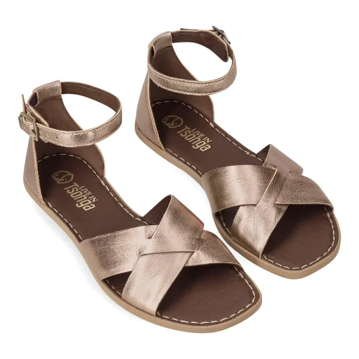 A pair of metallic rose gold flat sandals with criss-cross straps and ankle straps, shown on a plain white background.