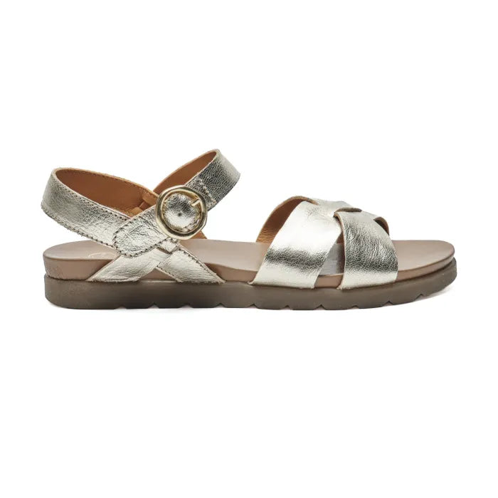A metallic silver leather flat sandal with crisscrossing straps, an adjustable ankle strap with a gold-tone buckle, and a brown contoured footbed, shown in profile against a white background.